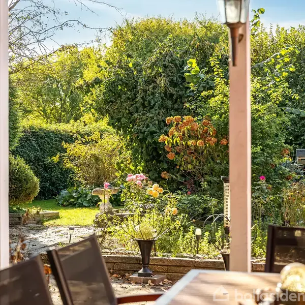 Terrasse møbleret med stole står stille under en pergola i en frodig have. Omkring er der blomsterbede og tæt buskads omgivet af sollys, hvilket skaber en hyggelig atmosfære.