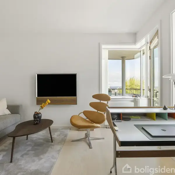 Stue med grå sofa, rektangulært sofabord med vase, moderne orange lænestol. Fladskærms-TV på væggen. Arbejdsbord ved vinduet med udsigt til balkon og natur.
