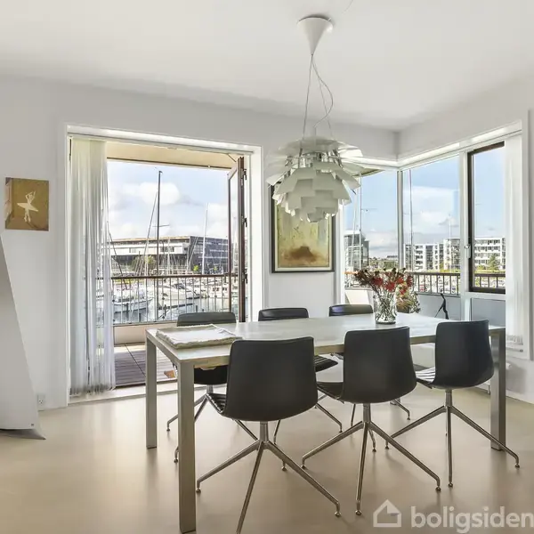 Spisebord omgivet af seks sorte stole står i et lyst rum med åbne døre til en balkon. Vaser med blomster pryder bordet. Kunstværker hænger på væggene.