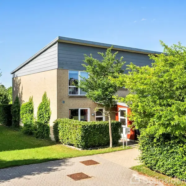 Et moderne hus med fladt tag og beige facade står i en grøn have. En indgang med røde detaljer er omgivet af hække og træer under en klar blå himmel.