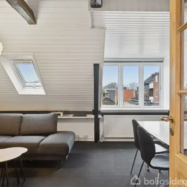 En grå sofa står ved et rundt træbord under en loftslampe, placeret i en lys stue med skråloft og store vinduer, der har udsigt til en bydel.