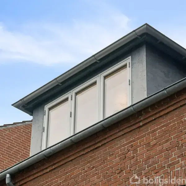 Et vindue er placeret på en tagkonstruktion af mursten. Vinduet, der er firkantet med tre sektioner, åbner udad mod en blå himmel.