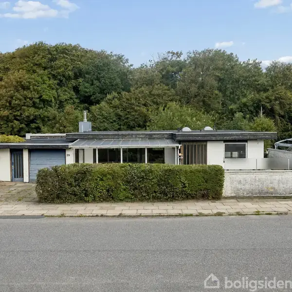En moderne, lav bungalow med fladt tag står stille mellem to huse, omgivet af en hæk. Baggrunden består af frodig skov. Adressen "Boligsiden" ses ikke i beskrivelsen.
