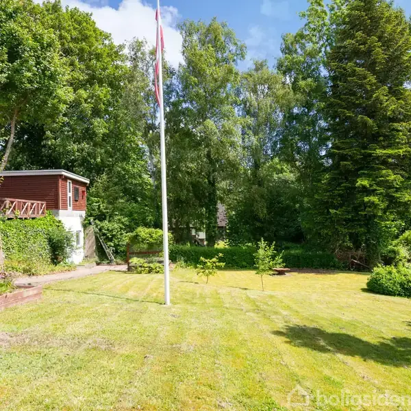 En stor have med en flagstang i midten. Omkranset af frodige træer og buske, der giver en grøn baggrund. En rød bygning med terrasse er på venstre side.
