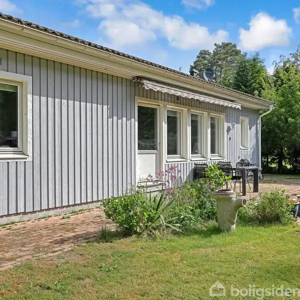 Et gråt træhus står stille i en have med græsplæne og blomsterbed. Ved siden af huset ses en terrasse med møbler og omgivet af buske og træer.