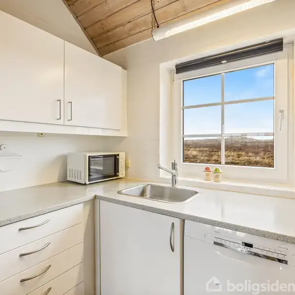 Køkken med hvide skabe og skuffer, kaffemaskine og mikrobølgeovn på bordet, udsigt til natur gennem vinduet, lyse trævægge og loft, træskærebræt står op ad væggen.