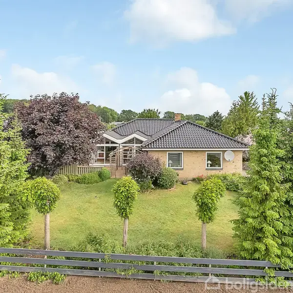 Et stuehus med mørkt tag omkranset af en velholdt have, fyldt med grønne buske og træer. Huset ligger i et beboelseskvarter med nærliggende huse og blå himmel.