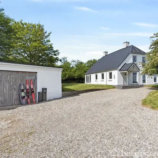 Et hvidt hus med sort tag står ved siden af en grusvej. Til venstre ses en garage med trædør og surfbrætter op ad væggen. Omkring er der grønne træer og græs.