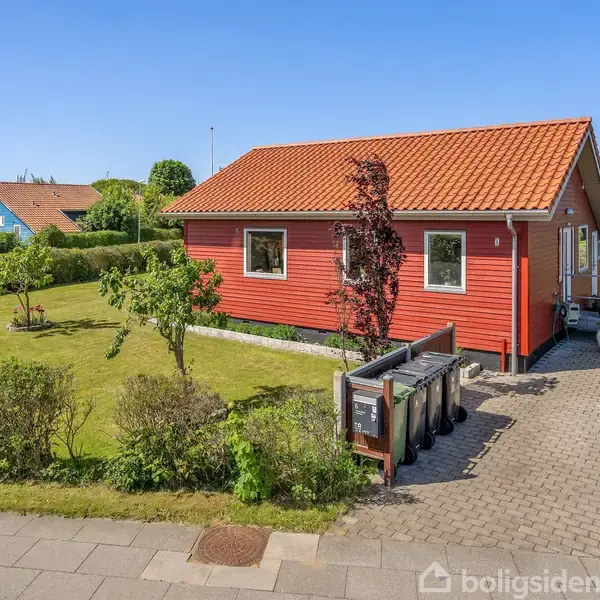 Et rødt hus med tegltag står på en velplejet græsplæne. Huset har vinduer og en indgangsvej med fliser. Haven omkranses af buske og små træer i et roligt boligområde.