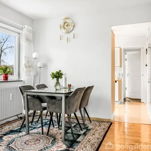 Et spisebord med fire stole står på et mønstret tæppe, omgivet af hvide vægge. Et vindue med gardiner lader lys ind, og der er grønne planter på bordet og vindueskarmen.