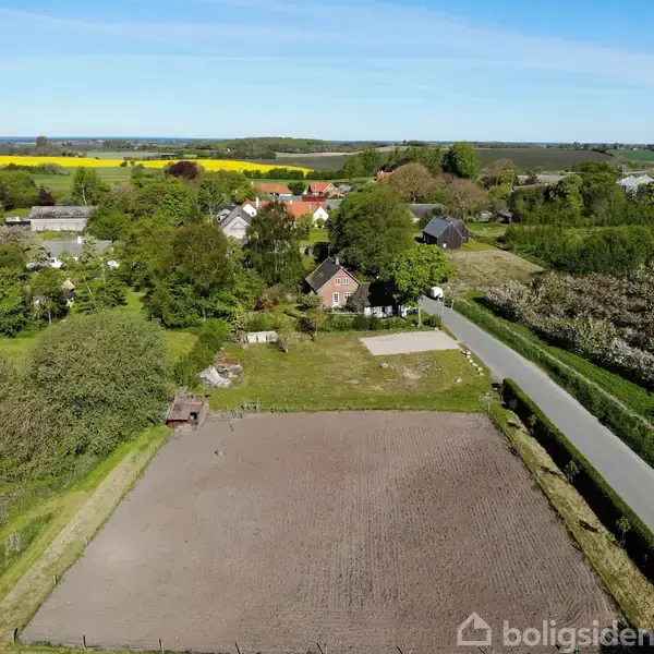 En mark ligger ved siden af en vej nær en landsby. Omkringliggende huse er omgivet af træer og marker. Landskabet er landligt med åbne marker og en blå himmel.