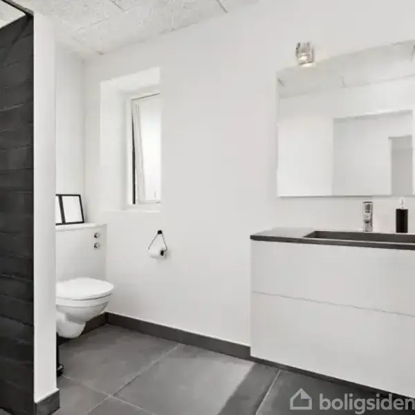 Et moderne badeværelse med en hvid håndvask på en skabsenhed til højre, et stort spejl ovenfor. Til venstre er der en toilet og en bruseniche med sorte fliser.