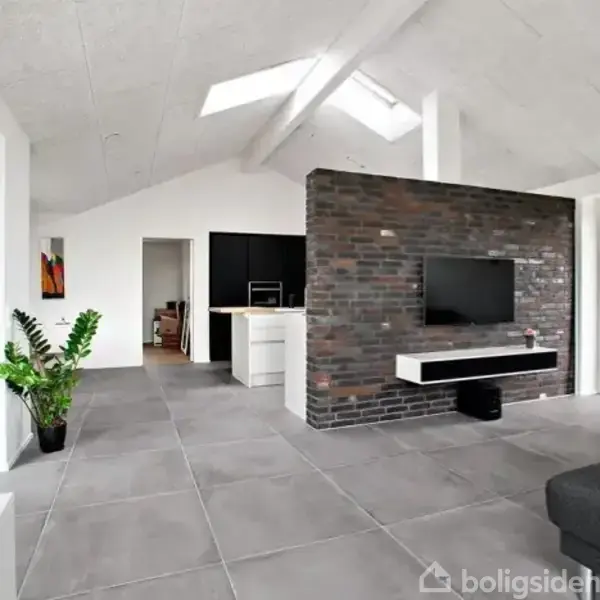 Stue med gråt klinkegulv indeholder en sort tv-væg i mursten, fladskærms-tv, og sort sofa. Planter og kunst beriger rummet, mens lys strømmer ind fra ovenlysvinduer.
