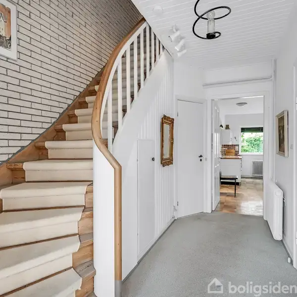 En trætrappe fører opad i en lys gang med hvidmalede vægge. I konteksten ses døre og en murstensvæg med et maleri. Belysning hænger i loftet.