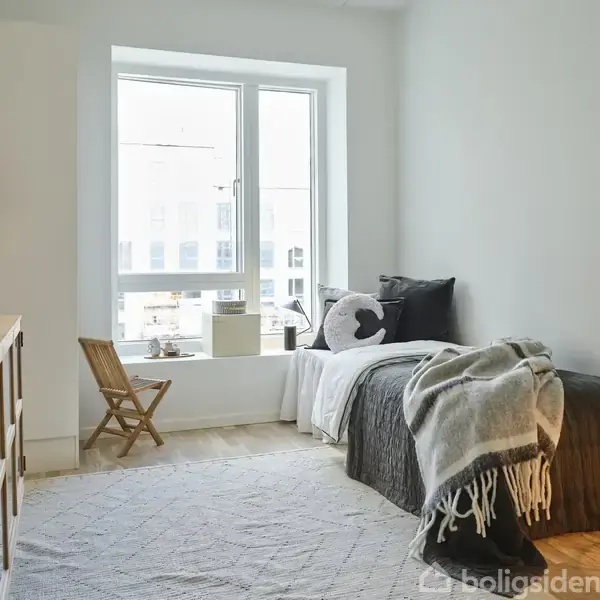 Seng dækket med gråt sengetøj står ved vindue. En træstol ved siden af. En lampe lyser op på et bord. Rummet er lyst og minimalistisk indrettet.