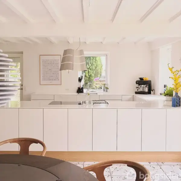 Køkkenø i moderne køkken, omgivet af hvide skabe, keramisk kogeplade og stor hængelampe. Vinduer viser udsigt til have. Vase med gule blomster står på bordplade.