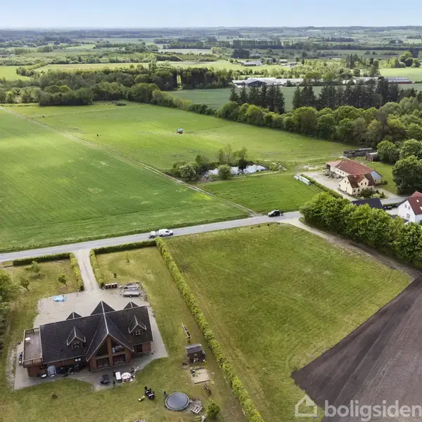 Hus ved kanten af en mark med grønne marker og skov i baggrunden. Andre huse og en vej ses i nærheden, omgivet af frodige græsarealer og marker.