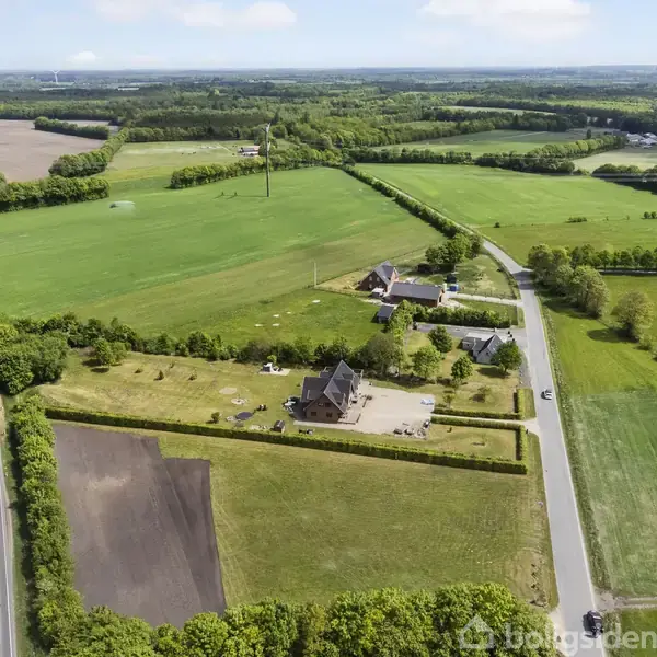 En gård ligger midt i landbrugslandskab, omgivet af marker og træer. Veje løber langs ejendommen, der også indeholder en stor have med græsplæne.