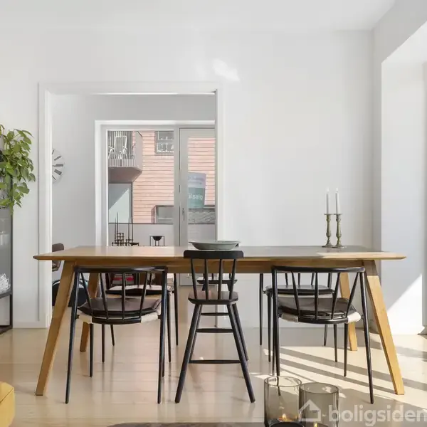 Et spisebord med sorte stole står foran en glasdør, der fører til en terrasse. Til venstre en vitrine fyldt med glas og en plante.