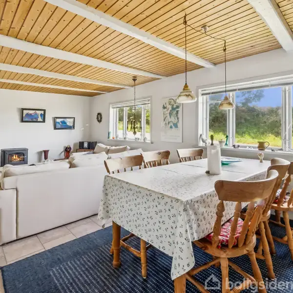 Stue med hvid sofa og pejs, bord dækket med dug omgivet af træstole. Store vinduer giver udsigt til naturen. Træloft og lamper skaber en hyggelig atmosfære.