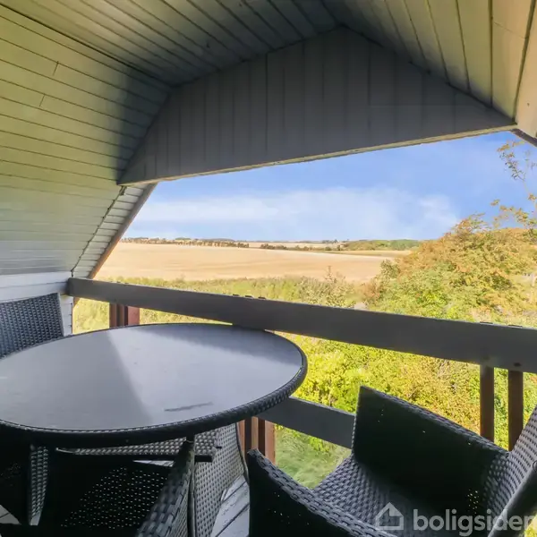 Et rundt bord omgivet af fire flettede stole står på en overdækket altan. I baggrunden ses åbne marker og blå himmel, hvilket skaber en naturskøn udsigt.