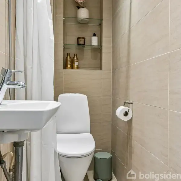 Et lille badeværelse med toilet og håndvask, der er adskilt af et forhæng. Hylder i baggrunden med flasker og dekorationer. Toiletrulleholder ved væggen. Beige vægge.