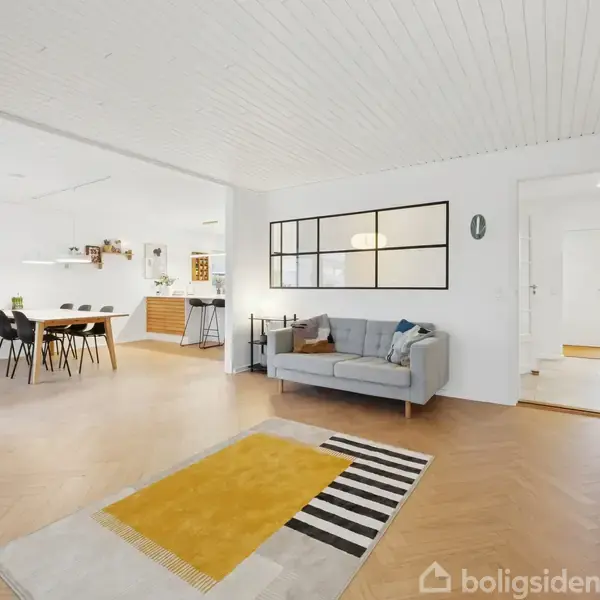 En moderne stue med en grå sofa står i forgrunden. Et gulvtæppe med gule og sorte striber ligger på trægulvet. Spisebord med stole ses i baggrunden.