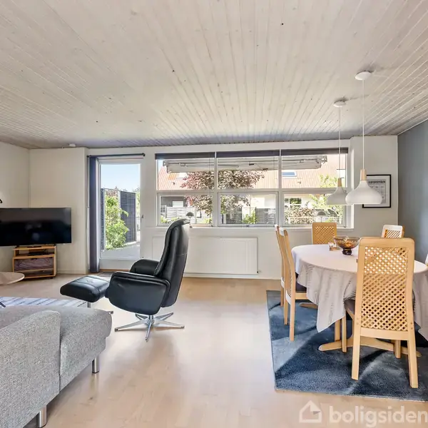 Stue med spisebord dækket med dug og stole på tæppe. Til venstre står en lænestol foran et stort vindue, fladskærms-tv på væggen og sofa. Indretning er lys og moderne.