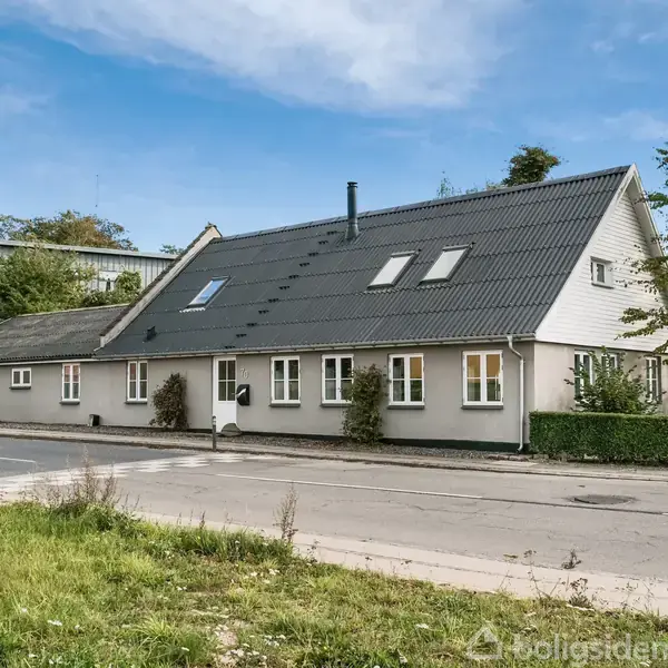 Hus med gråt skråtag har flere vinduer med hvid ramme. Det står langs en vej omgivet af grønne buske og træer under en blå himmel.