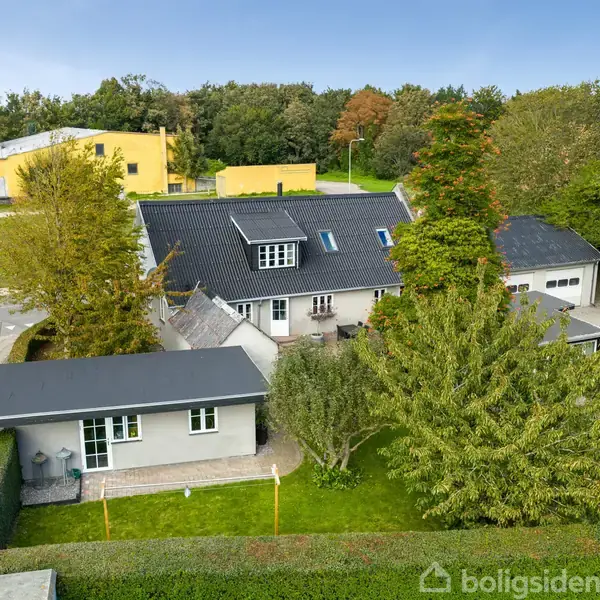 Hus med mørkt tag står stille omgivet af en frodig have og høj hæk. Sidevejen løber langs huset, mens der er flere omkringliggende bygninger og træer i baggrunden.