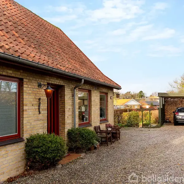 Murstenshus med rødt tegltag står ved en grusvej, hvor en bil er parkeret under en carport. Omkring huset ses hække og få buske.