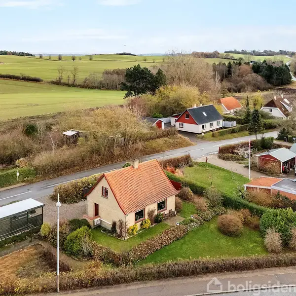 Et rødt tegltagshus står i en grøn have med buske. Det ligger i en landsby med flere små huse, omgivet af marker og træer under en klar himmel.