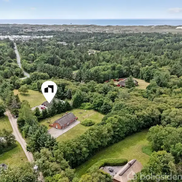 Et hus markeret med en hvid pind ligger i et grønt, skovklædt landskab omgivet af flere små bygninger og snoede veje. I baggrunden ses kystlinjen og havet.