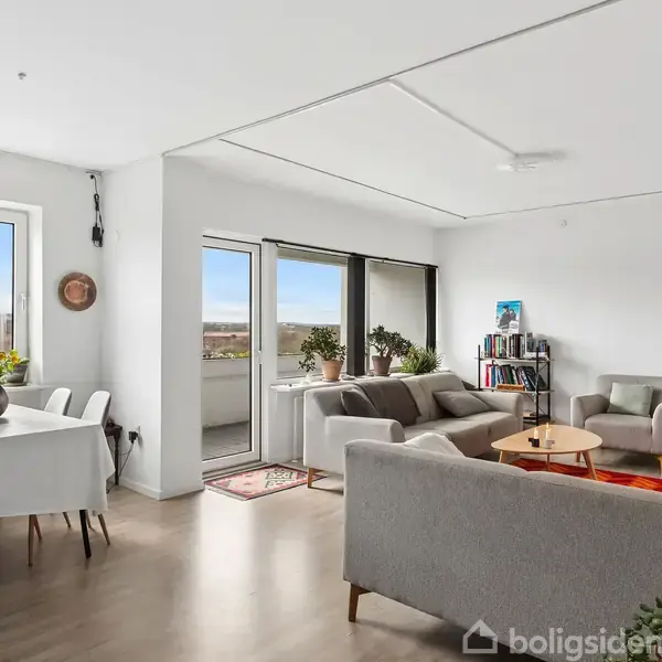 Stue med sofaer og sofabord, placeres i et lyst rum med store vinduer. Ved siden af er et spisebord med stole. Udsigt over landskab og balkon synlig udenfor.