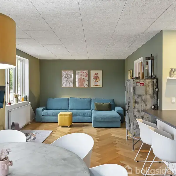 Blå sofa står i stue med lysegrønt væg, flankeres af gul skammel. Spisebord med hvide stole i forgrund, moderne køkken til højre. Billeder hænger over sofaen.