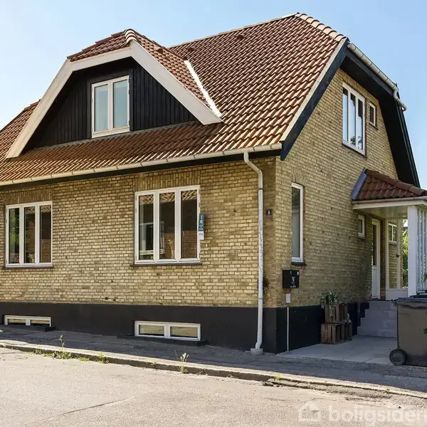 En murstensvilla med brune tagsten står på en stille gade. Huset har flere vinduer og en lille veranda. Det er omgivet af en have med træer og buske.