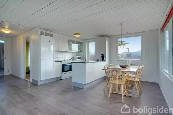 Et lyst køkken-alrum med hvidt køkkenudstyr og træmøbler. En spisebordssæt står ved et vindue med udsigt. Lysindfald fra flere vinduer giver rummet en hyggelig stemning.