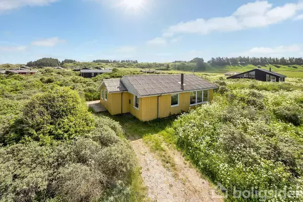 Et gult træhus står stille omgivet af frodigt grønt buskads på en solrig dag. Flere sommerhuse er synlige i det fjerne, under en klar, blå himmel.