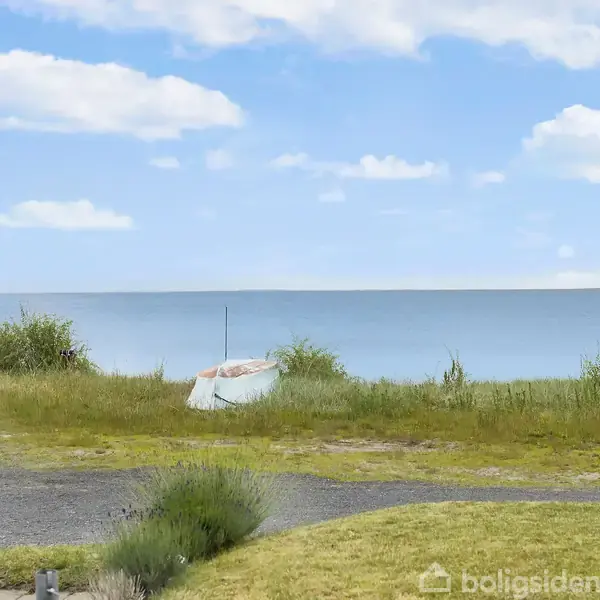 En lille båd ligger stille på en græsmark tæt ved havet under en blå himmel med spredte hvide skyer.