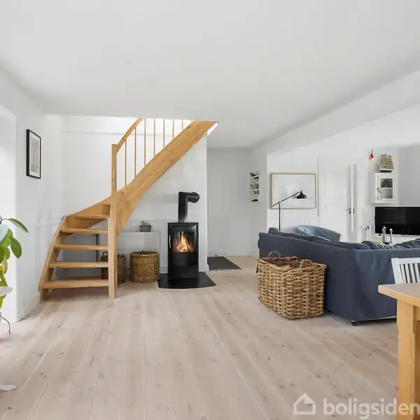 Trætrappe stiger ved siden af en kamin i et lyst stuerum; grå sofa, kurv, planter, og spisebord tilføjer hygge i rummets moderne indretning.