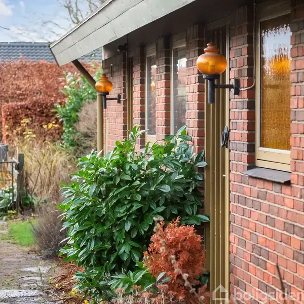 Rød murstensbygning med orange udendørs lamper hængende på væggen. En havegang flankeres af grønne buske og træer. Baggrunden viser en anden bygning og en vinterhimmel.