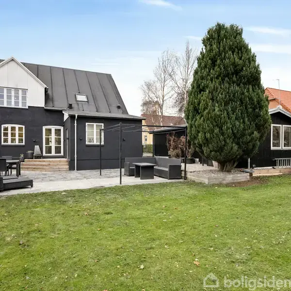 Sort hus med hvide vinduer står i en have med græs og stor terrasse. Et træ og en busk ses ved siden af, mens vejret er klart.