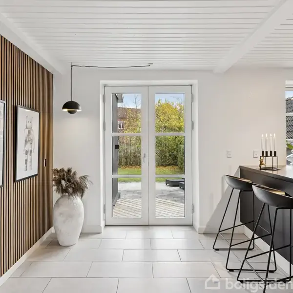 Dobbelt glasdør åbner ud mod en have; sort spisebord med barstole står til højre; til venstre hænger kunst på en væg med træpaneler.