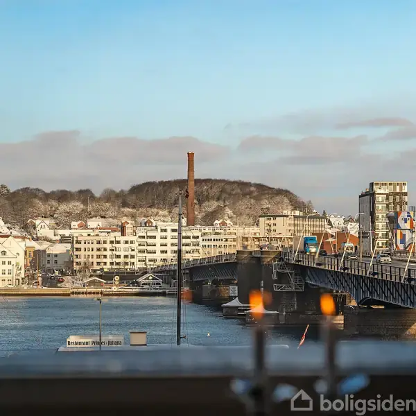 Bro strækker sig over vand i byområde, omgivet af bygninger og en bakke i baggrunden. Lys fra vindueskarm reflekteres i forgrunden.