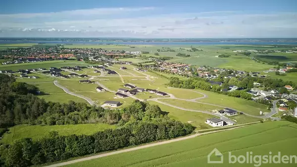 Luftfoto af et parcelhuskvarter under opførelse, omgivet af grønne marker og skovområder, med en by og kystlinje i baggrunden under en klar himmel.