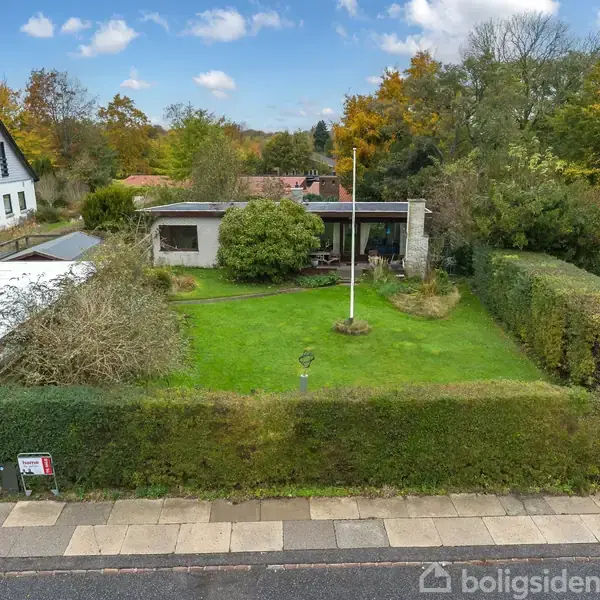 Et enkelt hus har en velplejet have med grønt græs og hække i forhaven. Et skilt ses ved hækken foran. Baggrunden viser flere træer og et andet hus.