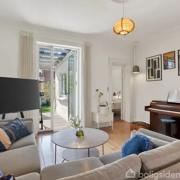 Stue med hjørnesofa og sofabord, vendt mod en stor fladskærm. Et klaver står ved siden af, og en dør åbner ud til en terrasse med udsigt til haven.
