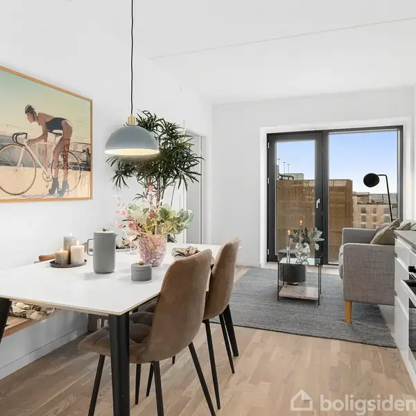 Spisebord med stole står i lys spisestue, pyntet med blomster og lys. Billedet på væggen viser en cyklist. Store vinduer fører til balkon med byudsigt.