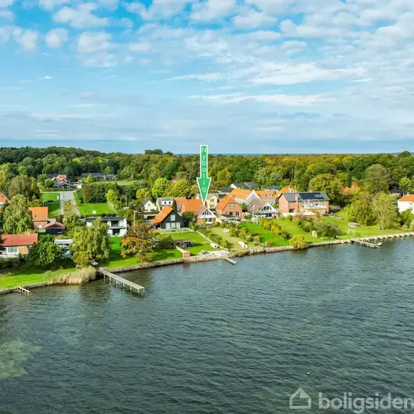 Hus markeret med en grøn pil ligger ved vandet, omgivet af flere huse og frodig natur. Skoven ses i baggrunden, mens himlen er blå med nogle skyer.