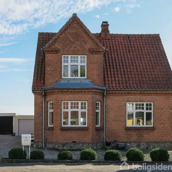 Et rødt murstenshus med rødt tegltag og flere vinduer. Det står langs en vej, omgivet af grus og buske. Himlen er skyet med blå nuancer.